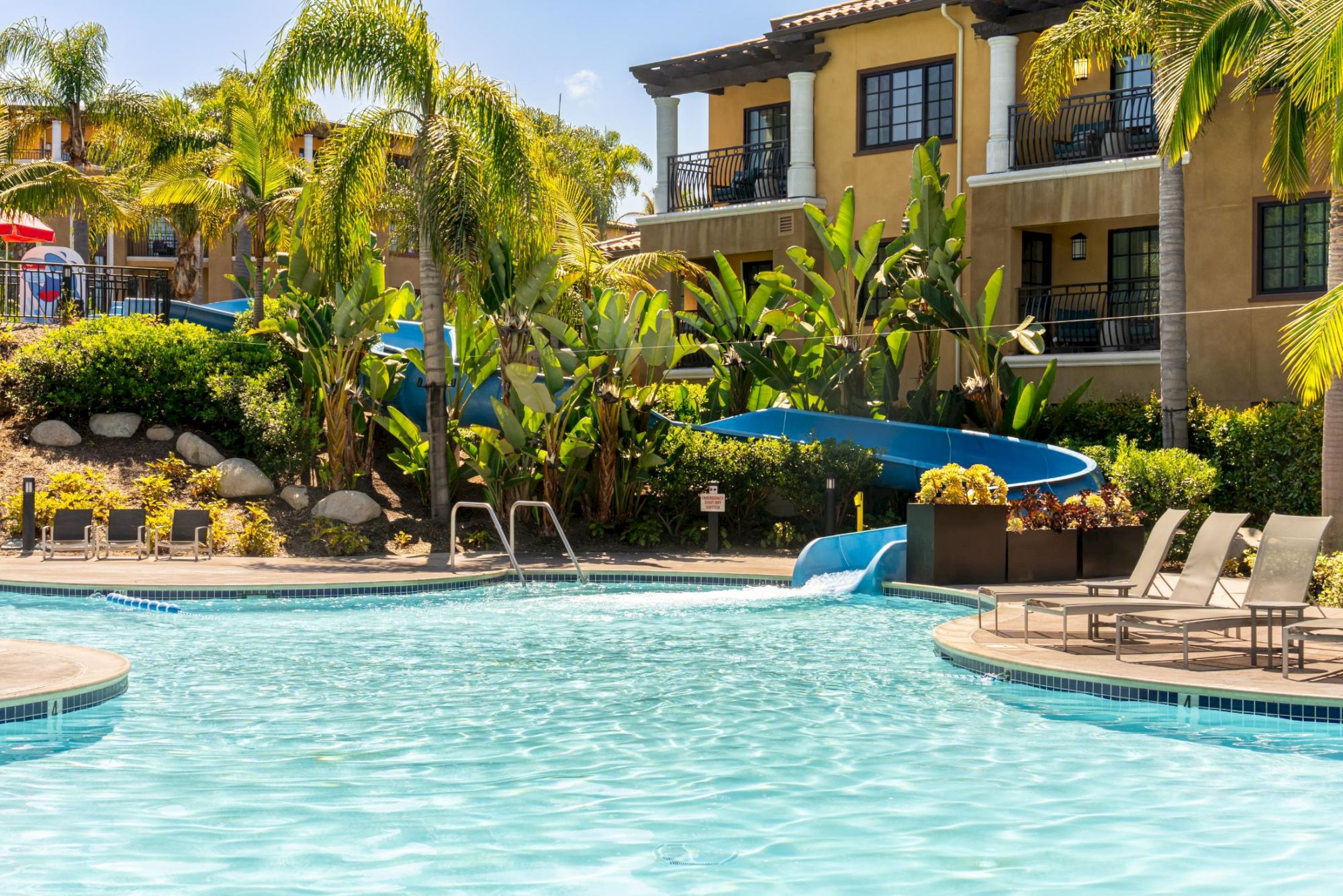Sunny hotel pool with a blue water slide, surrounded by palm trees, lush landscaping, and lounge chairs in front of a stucco building.