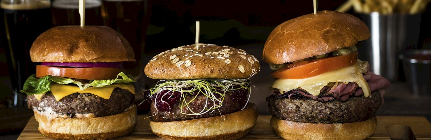 Three burgers on a wooden board, each with cheese and toppings, with toothpicks, like a caf&eacute; lunch lineup.