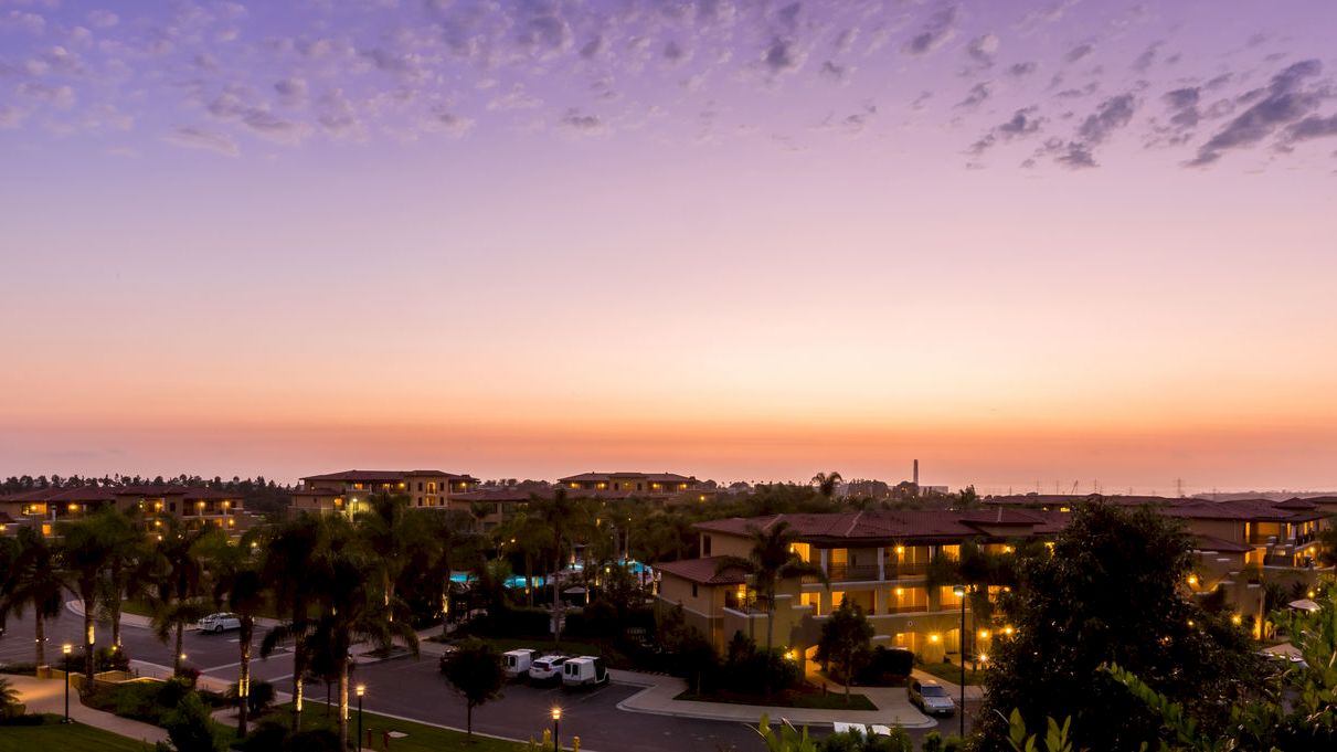 A purple sunset over a suburban neighborhood with palm trees, lit houses, and parked cars along winding roads.