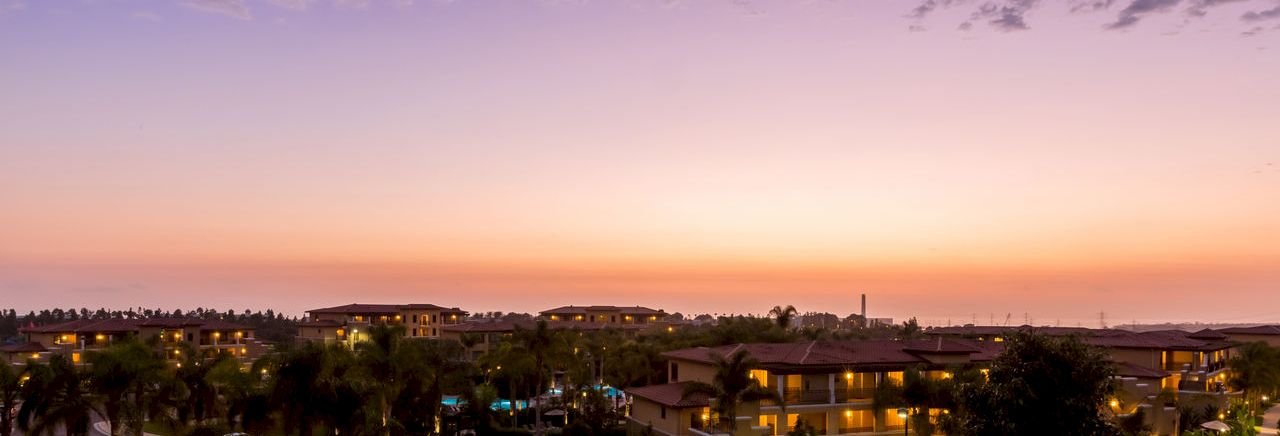 A purple sunset over a suburban neighborhood with palm trees, lit houses, and parked cars along winding roads.