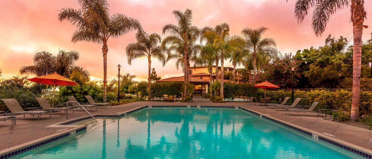 A turquoise hotel swimming pool at sunset, surrounded by palm trees, lounge chairs, and red umbrellas, under pink-orange clouds.