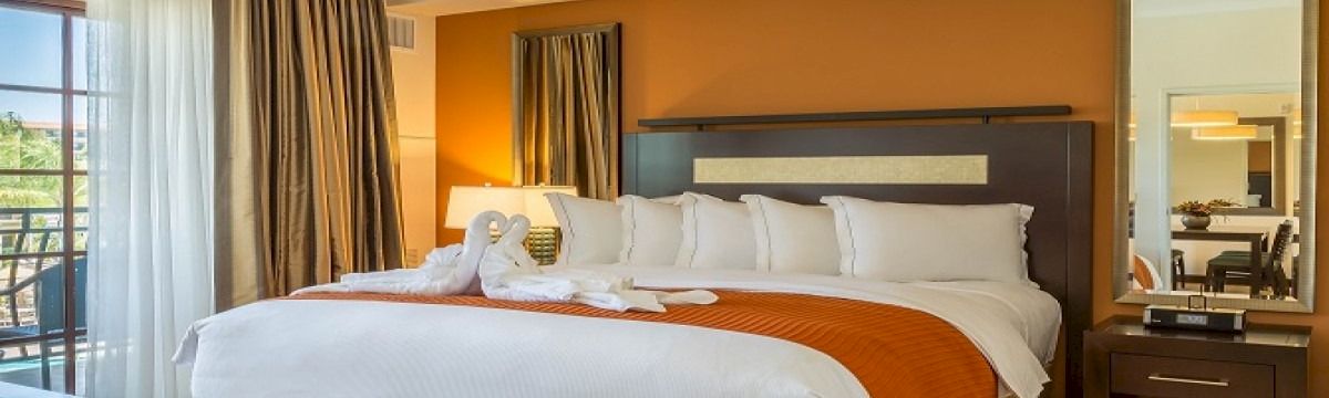 A well-made hotel room with a large bed, white linens, orange throw, wooden headboard, nightstands, mirror, and a balcony view.