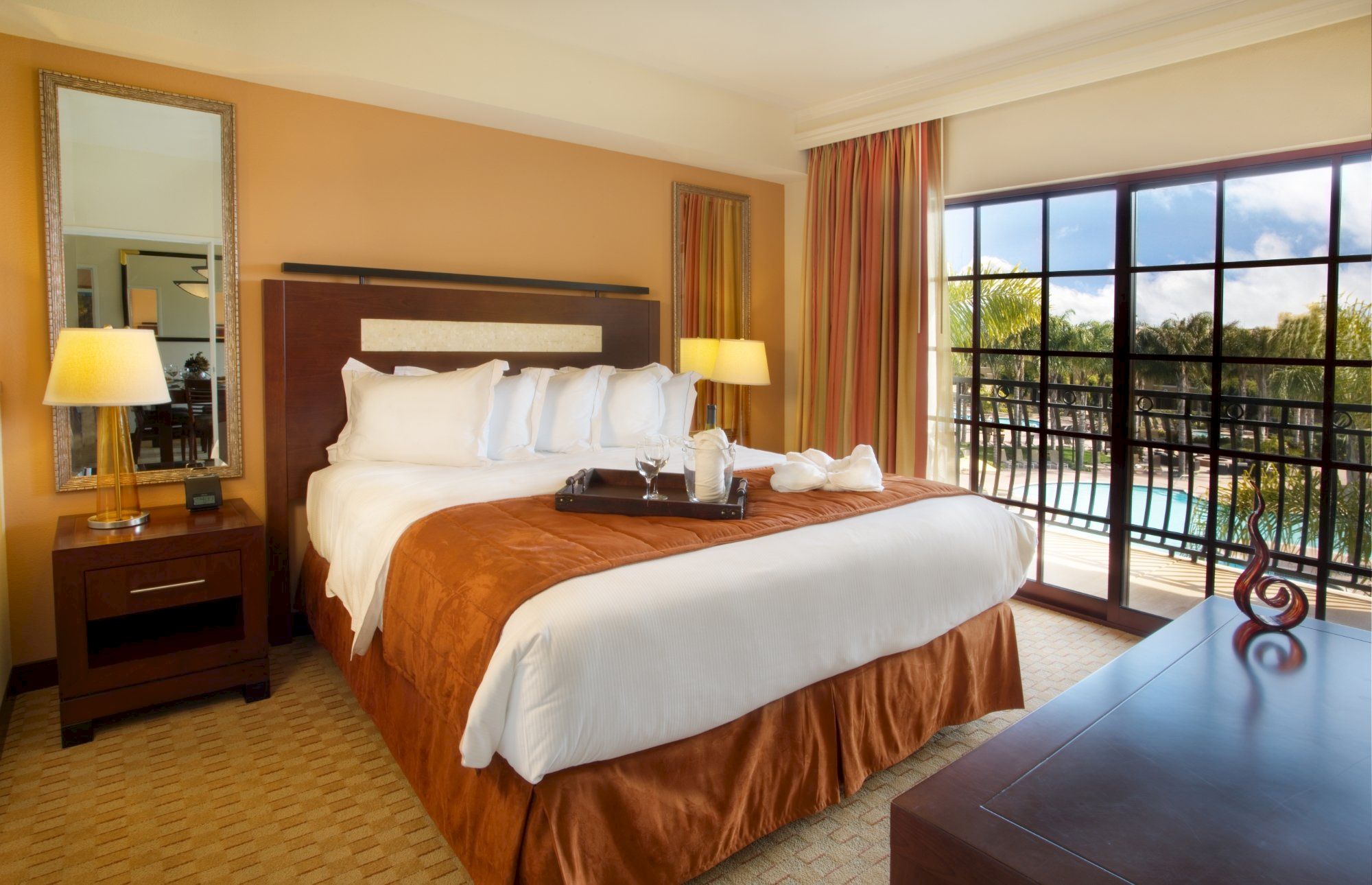 A bright hotel bedroom with a king bed, bedside lamps, and large windows overlooking palm trees.