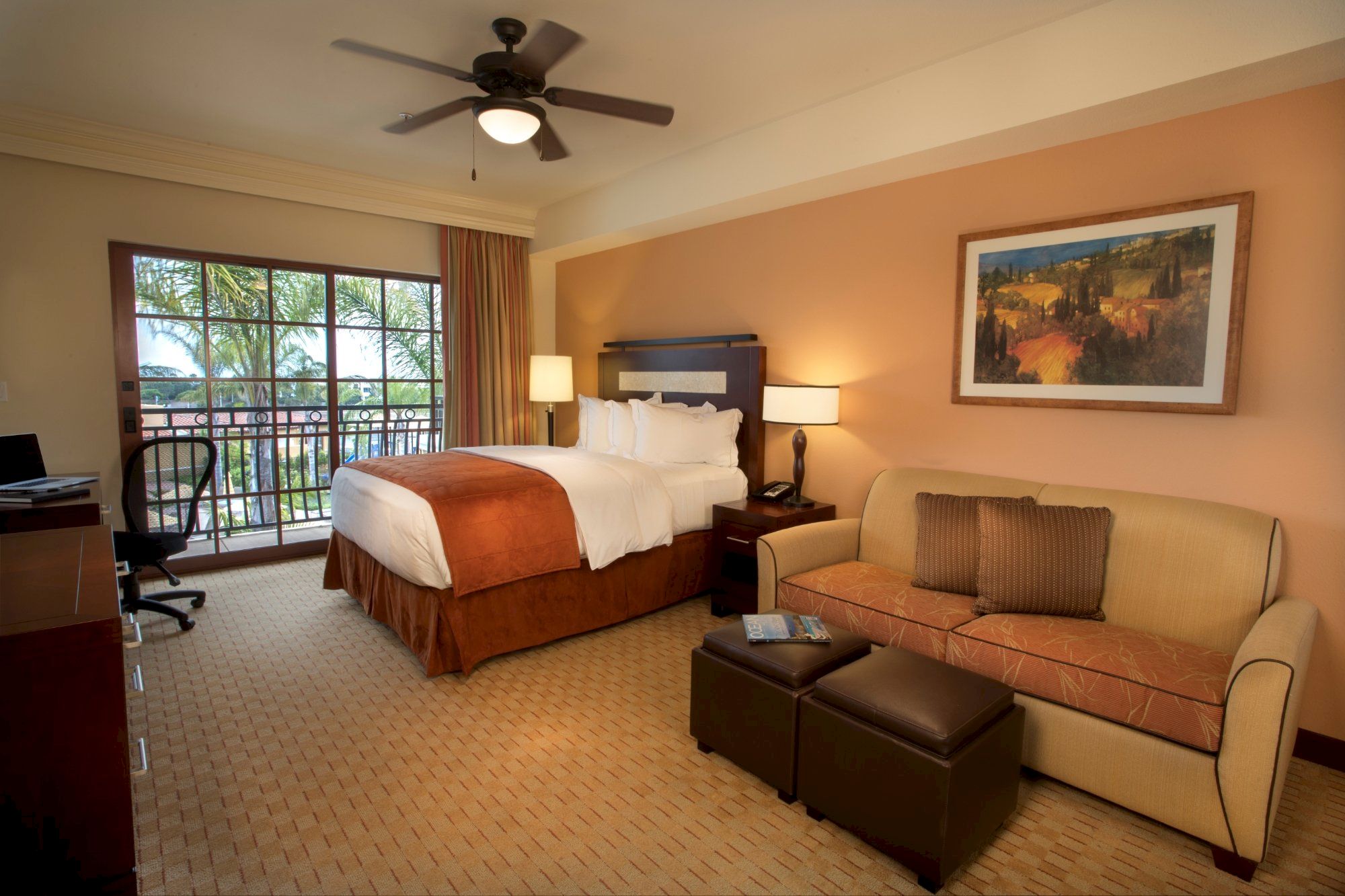 A hotel bedroom with a king bed, ceiling fan light, sofa, lamps, framed wall art, and large bay windows outside.