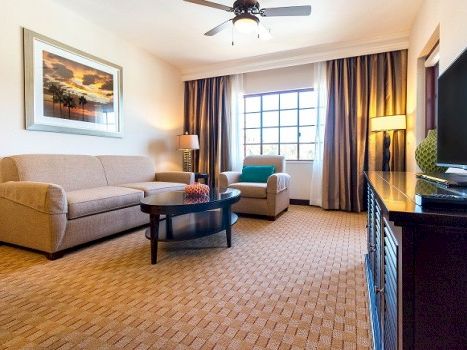 A cozy hotel living room with beige sofa, armchairs, coffee table, art on the wall, and a flat-screen TV across from a dark wood cabinet, warm lighting.
