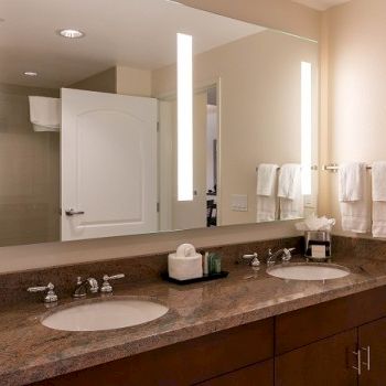 A spacious bathroom vanity with a double sink, earth-toned granite countertop, large mirror, wall-mounted lights, and neatly hung towels on the right.