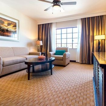 Cozy hotel living room with beige sofa, armchairs, round coffee table, warm lamps, large window, framed art, and a flat-screen on a dark cabinet.
