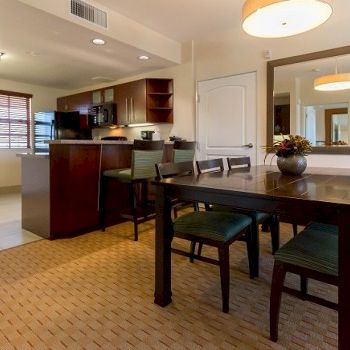 A cozy dining area with a dark wood table and six green-cushioned chairs, adjacent to a modern kitchen with a breakfast bar and entryway.