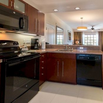 A modern open-plan kitchen with dark wood cabinets, black appliances, a breakfast bar, and a view into a cozy living area with a ceiling fan and natural light.