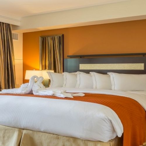 A bright hotel room with a large bed, white linens, orange throw, two nightstands, a window with curtains, and a reflection in a tall mirror.