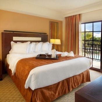 A neatly made hotel room with a large bed, matching nightstands, lamps, a balcony with a view, and warm earthy tones throughout.