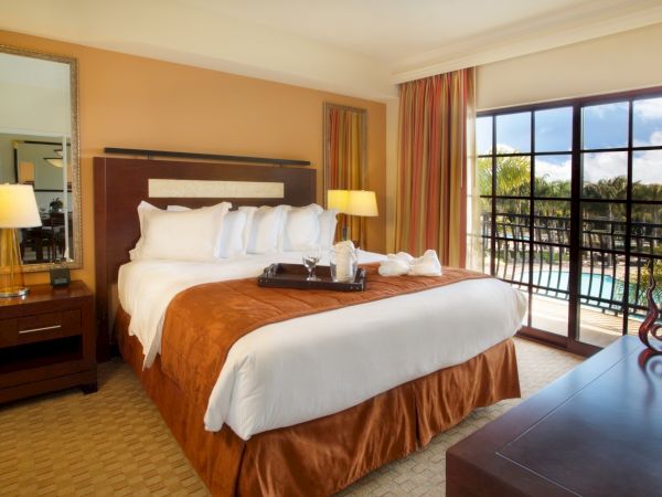 A tidy hotel room with a king bed, brown and white bedding, two nightstands with lamps, a large window/door to a balcony, and a dresser.