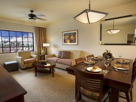 Cozy hotel suite with a living area, dining table set for four, large window, ceiling fans, and warm neutral decor.