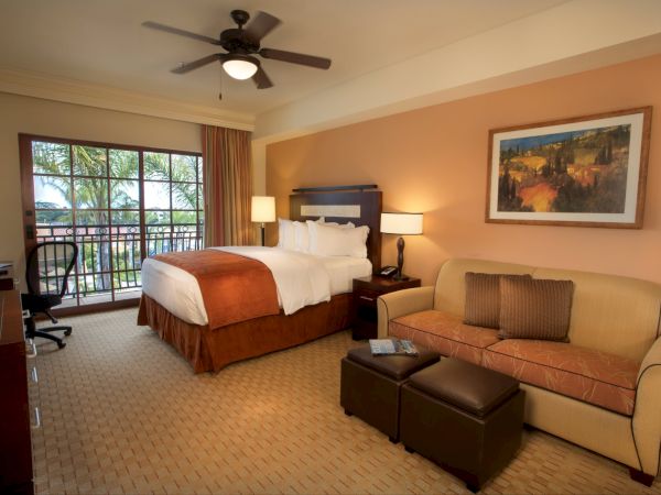 A hotel-style bedroom with a king bed, ceiling fan, two lamps, a sofa, large windows, and framed landscape art on the wall.