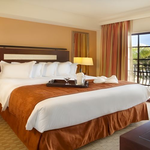 A hotel room with a large bed, brown-orange bedspread, two nightstands with lamps, a balcony with glass doors, a dresser, chair, and a mirror. The room looks bright and neat.