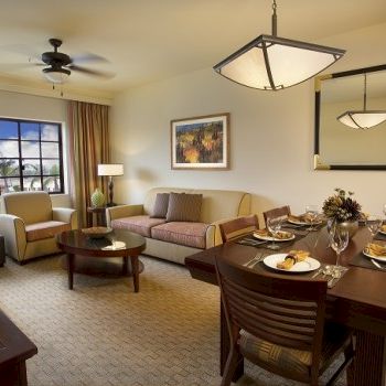 Cozy living and dining area in a hotel suite: sofa, chairs, coffee table, window view, and a dining table set for a meal, warm lighting.