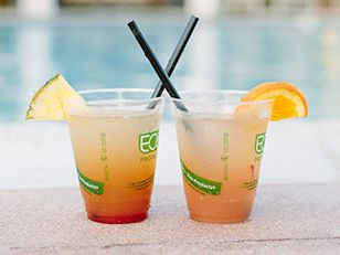 Two fruity drinks with lemon and orange wedges by a pool, each with a straw, plastic cups, and colorful layered juice.