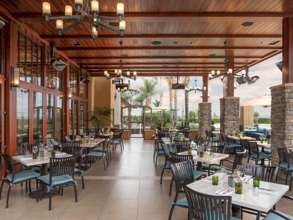 Open-air dining patio with neatly set tables, hanging lanterns, stone columns, wood ceiling, and glass walls, ready for a meal.