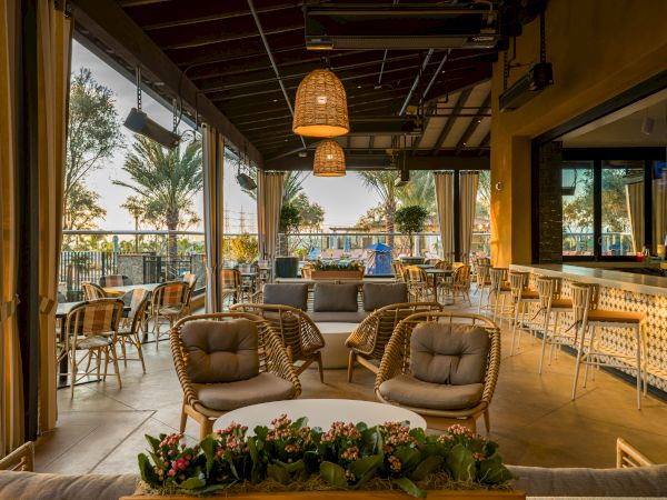 Warm, modern outdoor patio with wicker chairs, string-curtain drapes, hanging lamps, and a view of palm trees and a pool area, at sunset.