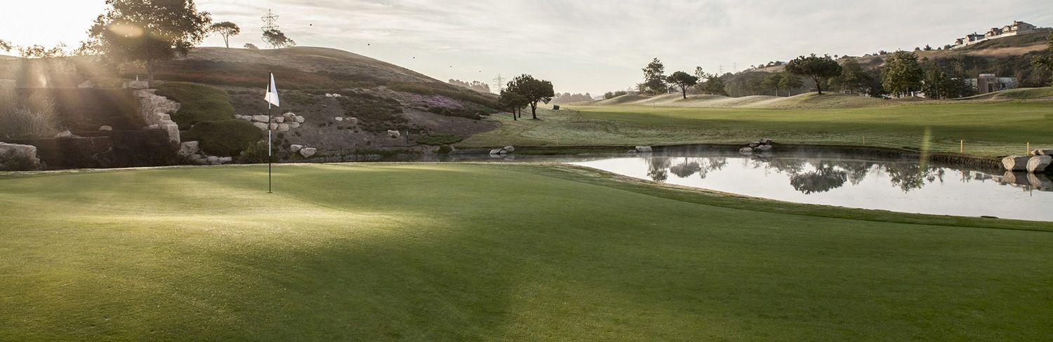 A sunny golf course fairway with a flag on the green and a calm pond reflecting hills and trees in the distance.