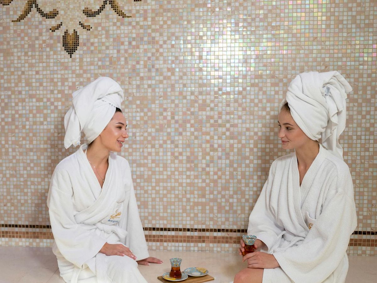 Two women in white bathrobes with towels on their heads sit in a spa, enjoying tea and conversation by a tiled wall.
