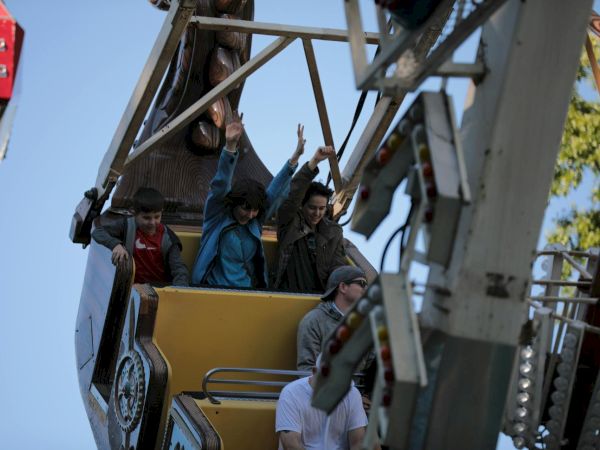 People riding a tall amusement park ride, waving and smiling from a yellow gondola as a safety harness clips in, with clear blue sky behind.