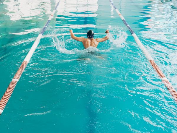 A swimmer dives into a bright indoor pool, arms outstretched, racing between lane ropes toward the far end.