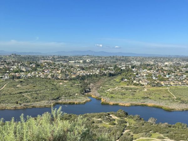 A scenic overlook shows a river winding through scrubby hills, with a valley of houses beyond and a clear blue sky above.