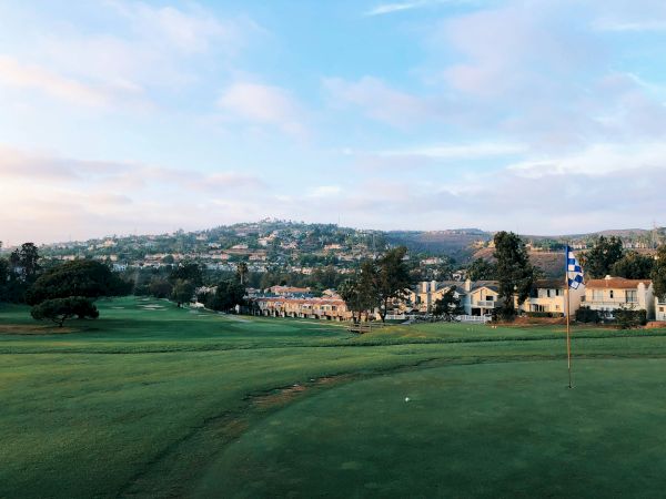 A serene golf course with green fairways, a flag on the green, and a quaint town with low hills in the background under a partly cloudy sky.