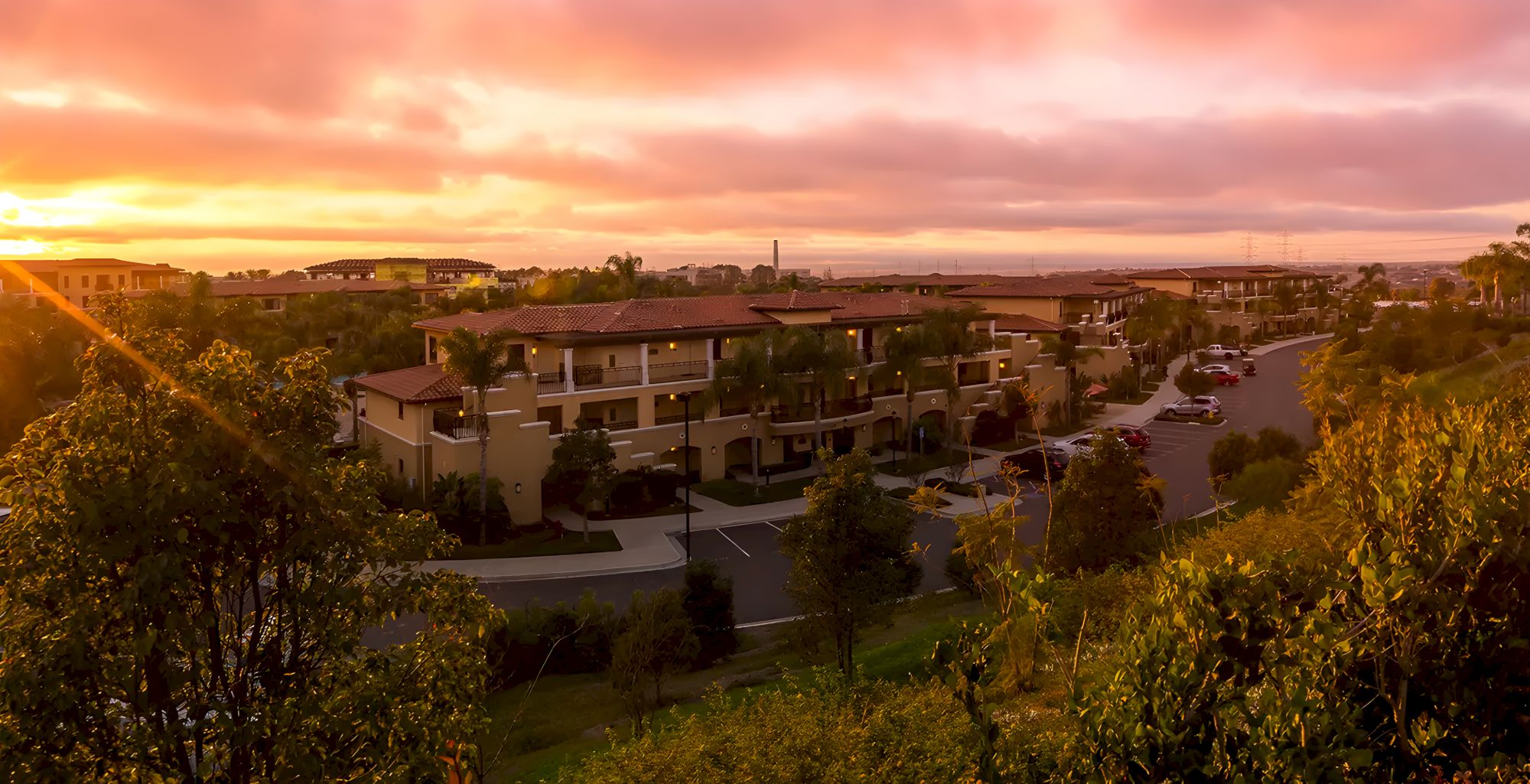 A quiet residential complex at sunset, with palm trees, parked cars, winding roads, and glowing orange clouds.