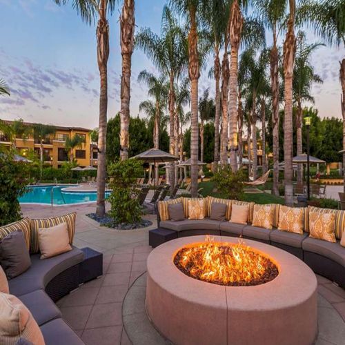 A stylish outdoor lounge with a circular fire pit, curved sectional seating, plush cushions, palm trees, and a swimming pool in the background.