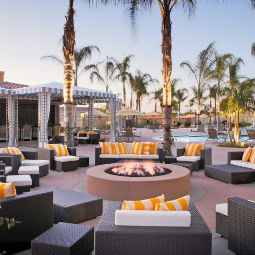 Sunset resort patio with a circular fire pit, striped lounge cushions, palm trees, and a swimming pool glinting in the background.
