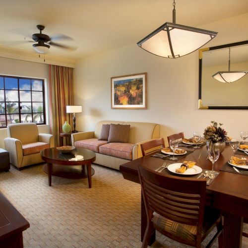 A bright living/dining room with a sofa, a glass dining table set for dinner, large windows, and hanging ceiling lamps.