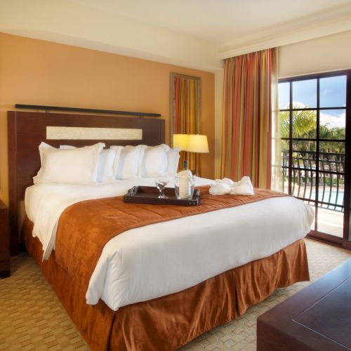 A cozy hotel bedroom with a large made bed, bedside lamps, and big windows overlooking palm trees outside.