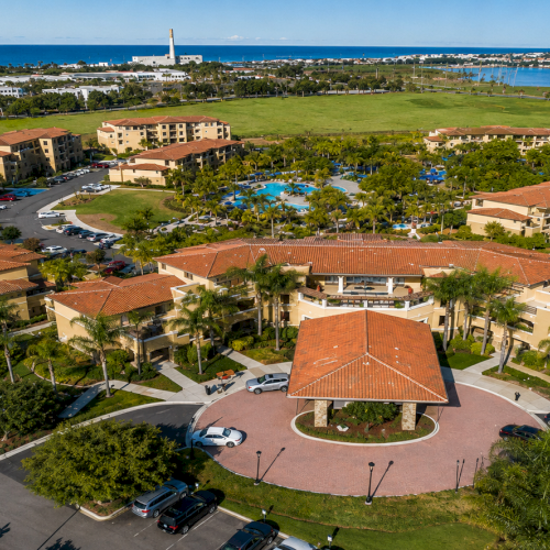 Aerial view of a seaside resort with terracotta-roof buildings, gardens, swimming pool, parking lots, and a calm blue ocean.