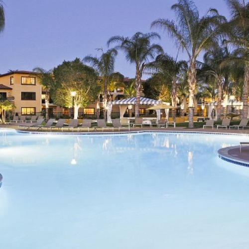 An outdoor resort swimming pool at dusk, lined with loungers and palm trees, with buildings in the background.