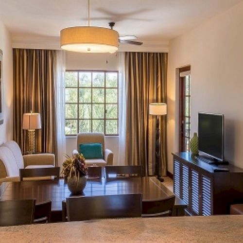 A bright hotel suite living area with sofa, armchairs, TV, coffee table, and large windows with dark curtains.