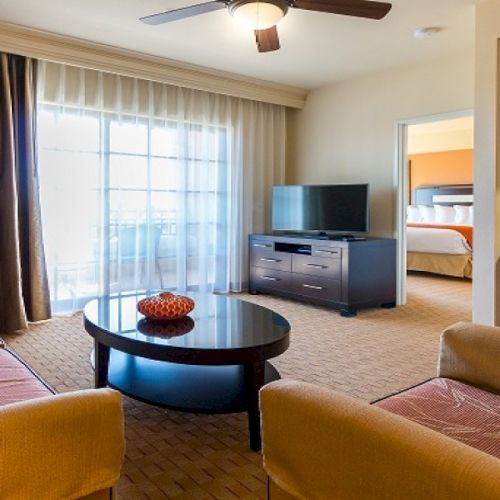 Bright hotel living room: sofas, round glass coffee table, TV stand, desk, and a bedroom visible through the doorway.