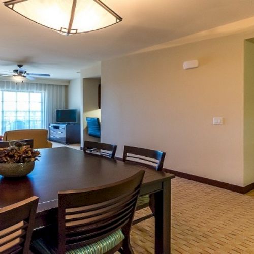 A bright apartment dining-living room with a table, chairs, TV, and a doorway leading to another room.