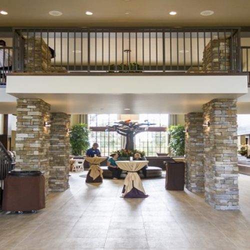 A modern hotel lobby with stone columns, stairs, and banquet-style tables with people setting up for an event.