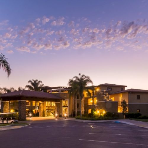An illuminated hotel or resort building at dusk, surrounded by tall palm trees and a quiet driveway with streetlights.
