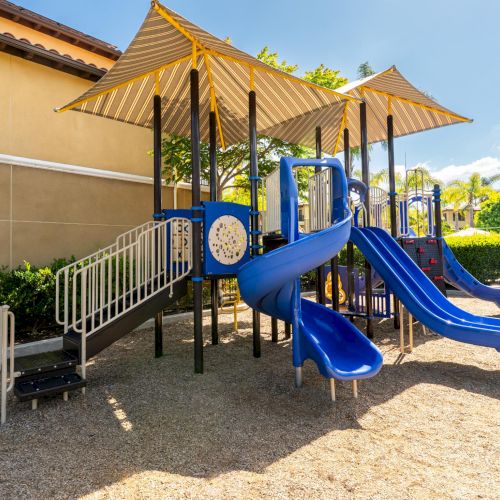 A sunny outdoor playground with blue slides, striped shade canopies, and railings by a building.
