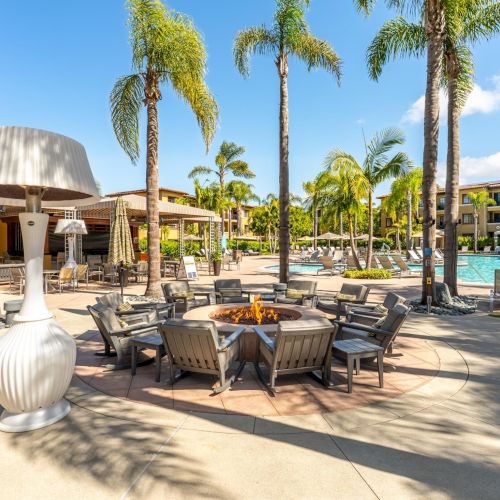 Sunny resort pool area with palm trees, lounge chairs, umbrellas, and a flaming fire pit by the water.