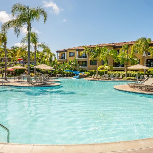 Sunny resort pool with palm trees, loungers, and a yellow apartment building under a clear blue sky.