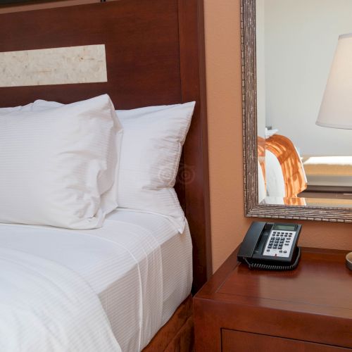 A hotel room shows a white bed, bedside table with an amber lamp, a phone, and a wall mirror reflecting the room.