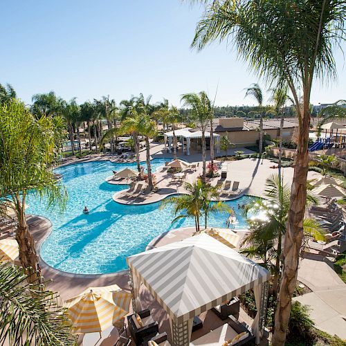 A sunny resort pool with loungers, striped umbrellas, palm trees, and a clear blue sky stretching over a landscaped courtyard.