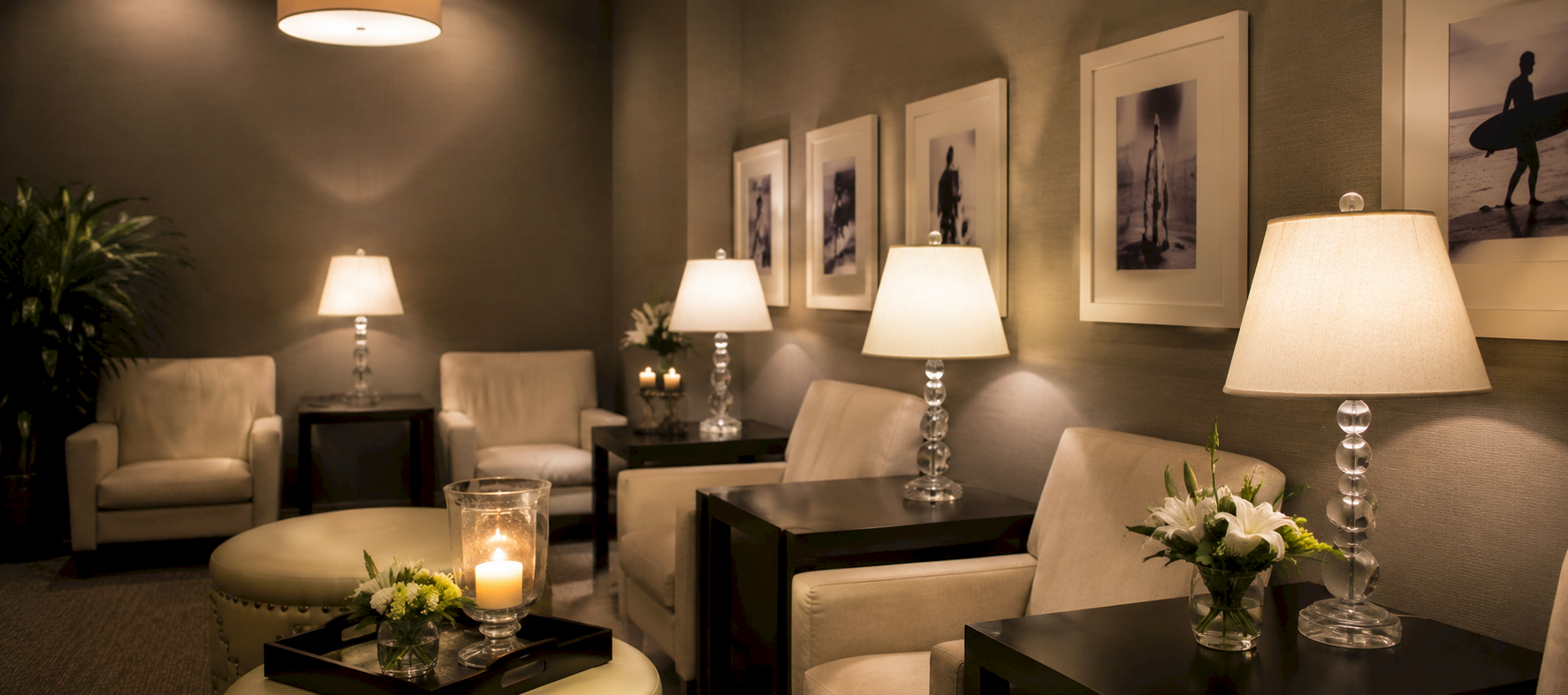 A cozy lounge with neutral seating, lamps glow warmly, framed photos on the wall, and small tables with candles and flowers.