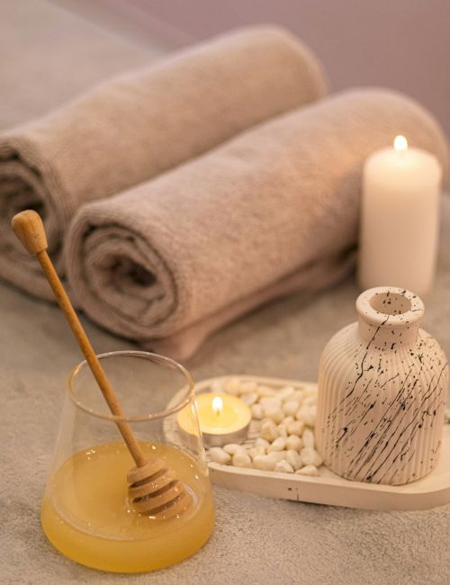 A cozy spa setup: rolled towels, a lit candle, a small jug and stones, plus a honey dipper resting in a yellow bowl&mdash;pure relaxation.