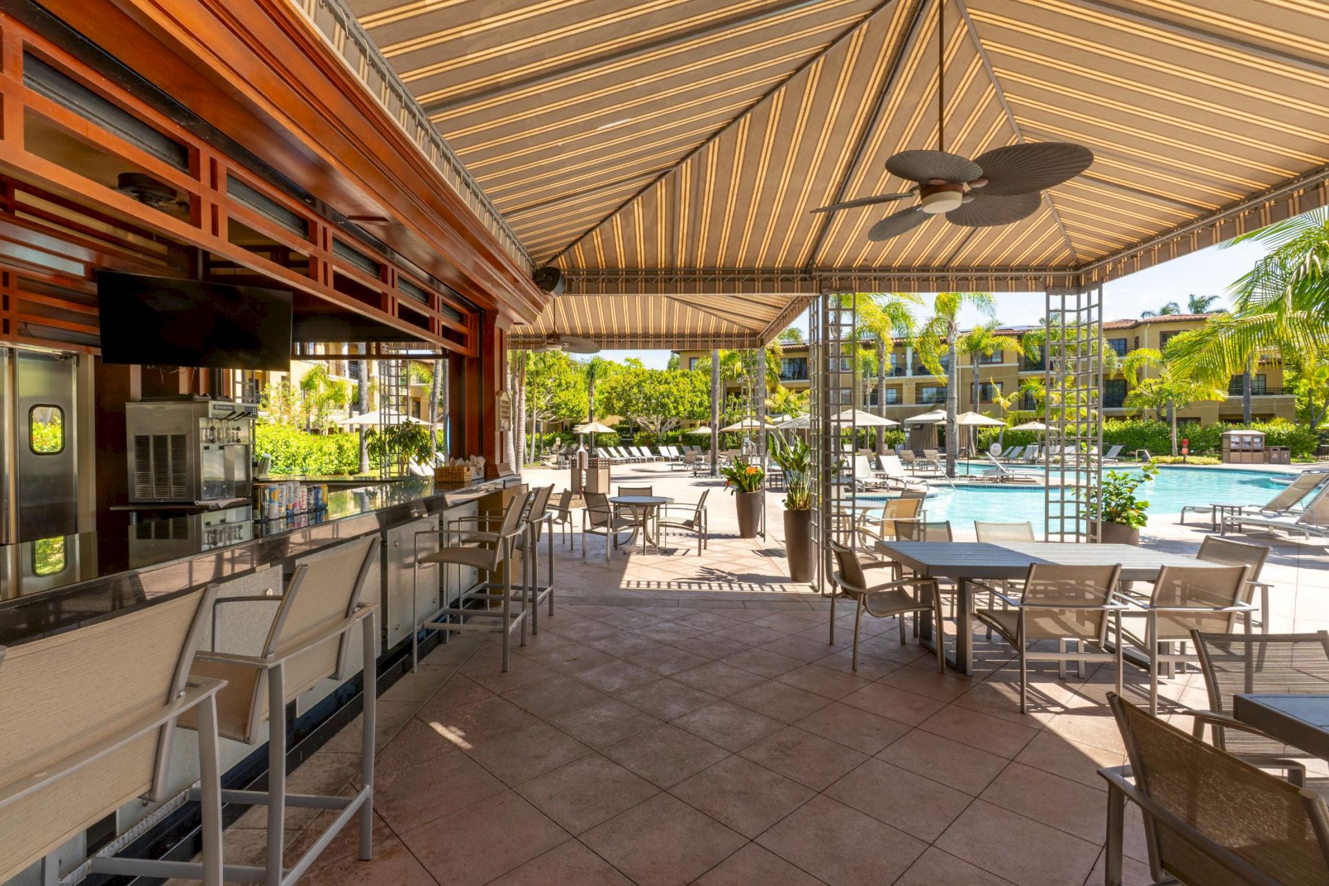 Resort poolside patio with lounge chairs and tables, plus a bar/ice setup under a striped canopy beside a swimming pool.